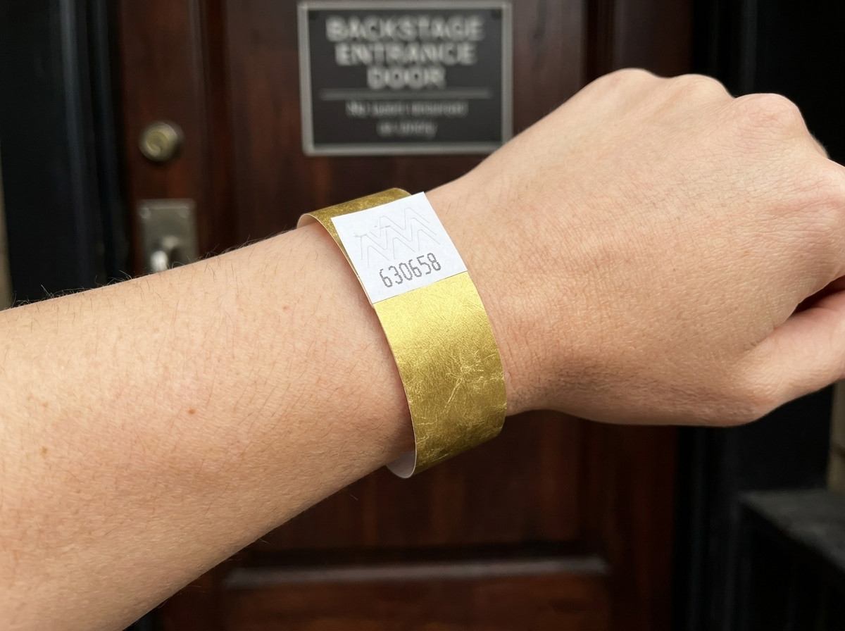 Pulseira VIP de festival com impressão metalizada dourada e codigo de barras, usada no pulso em frente a uma entrada backstage